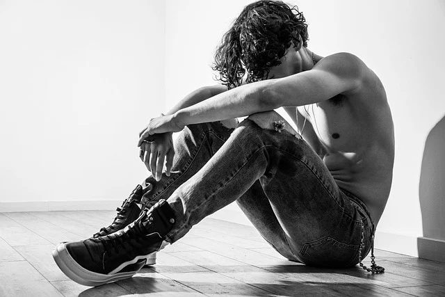 A shirtless man sitting on the floor wearing slim jeans and sneakers
