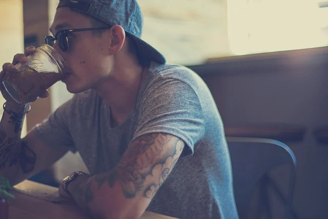 A man with tattoos wearing a cap and grey t-shirt drinking