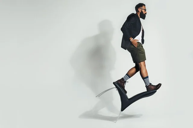 A man wearing a blazer, shorts, and boots standing on a tilted chair