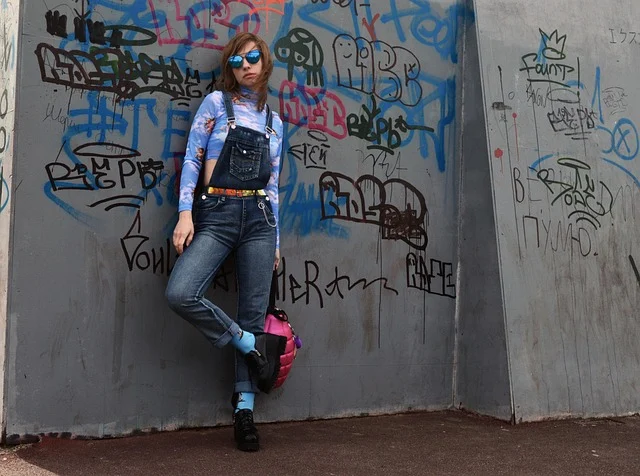 A woman wearing denim overalls leaning against a graffiti wall