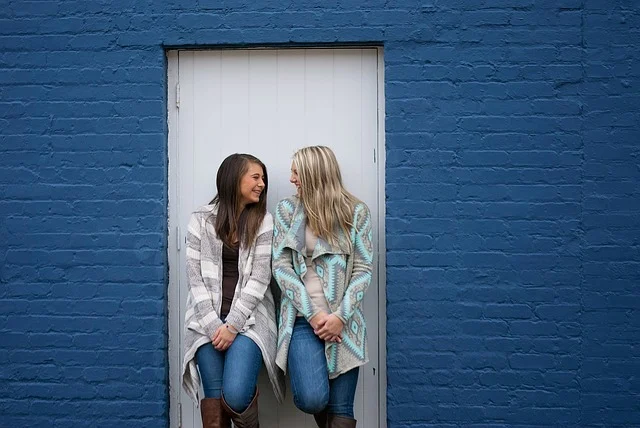 Two women wearing sweaters and jeans leaning against a door