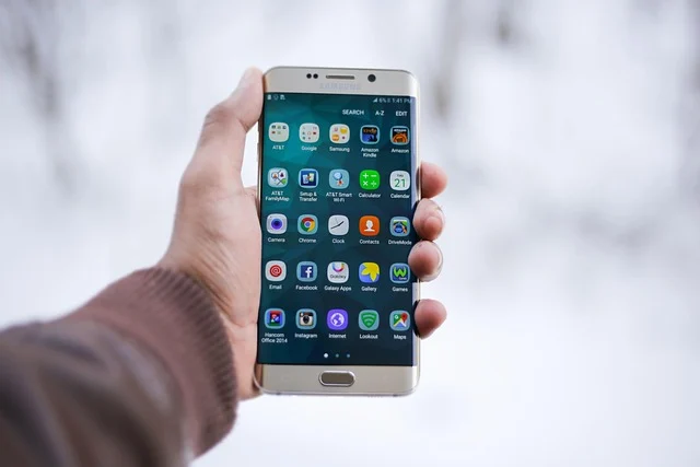 A hand holding a smartphone displaying various app icons outdoors