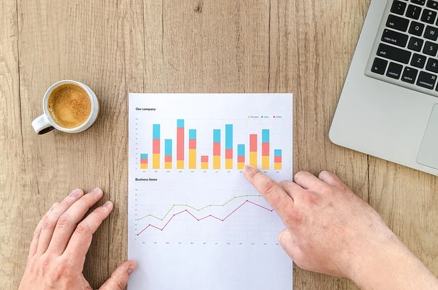 A person pointing at financial charts on a desk next to a laptop
