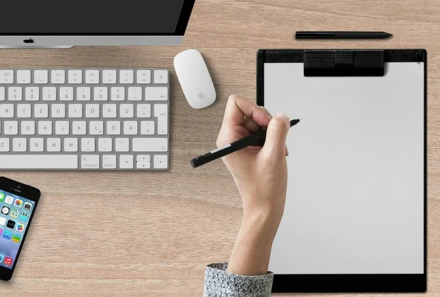 A hand writing on a blank clipboard near a keyboard and phone