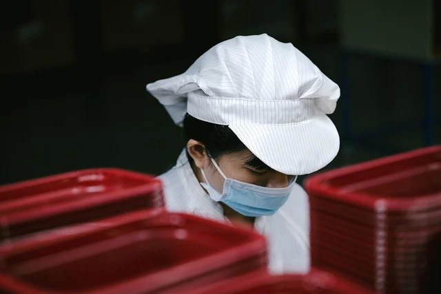 A factory worker wearing a cap and mask handling plastic containers