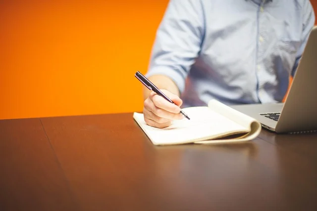 A person writing in a notebook next to a laptop