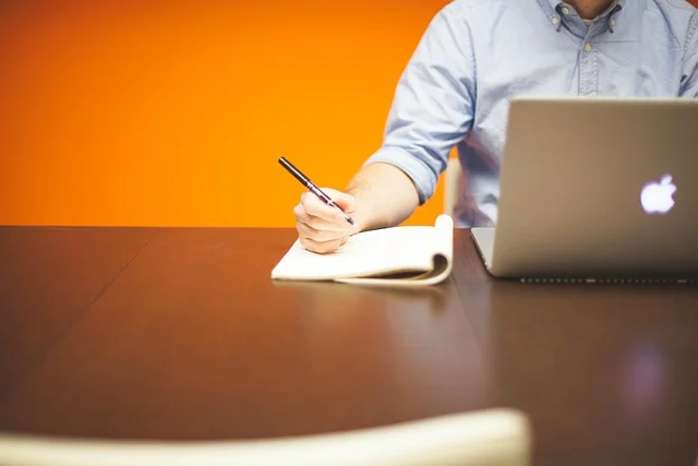 A person writing in a notebook next to a laptop