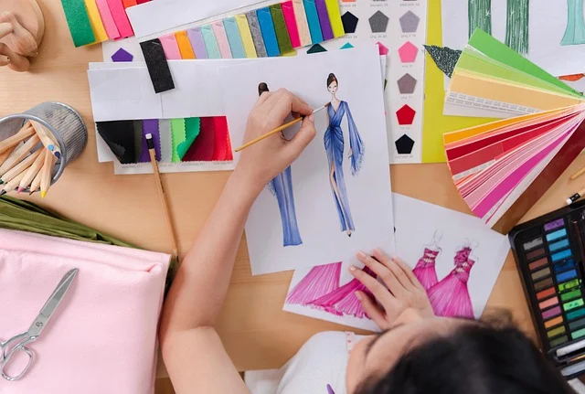 A fashion designer painting dress sketches surrounded by fabric swatches and art supplies