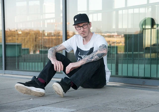 A tattooed man sitting outdoors wearing a graphic t-shirt and cap