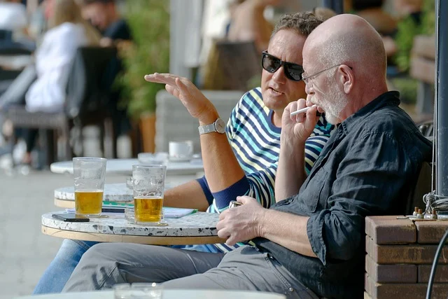 Two men at an outdoor cafe drinking beer and smoking a cigarette