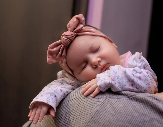 A sleeping baby with a pink headband resting on someone's shoulder
